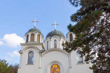 Sırbistan 'ın başkenti Belgrad' daki çarpıcı beyaz Crkva Sveti Jovan Vladimir 'in güneş ışığında görünen yüzü. Kubbeleri ve haçları yeşil çam ağaçlarıyla çevrili mavi gökyüzüne doğru uzanır..