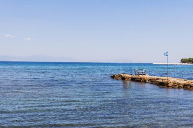 Berrak mavi suyla sakin deniz manzarası, dinlenmek için görkemli dağlara ve canlı kıyı bitkilerine karşı kurulmuş bir bank ve bayrak direğinin yer aldığı tuhaf kayalık iskele..