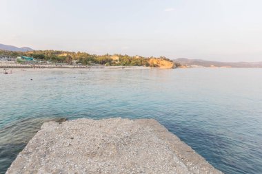 İnsanlarla, ağaçlarla ve binalarla dolu yoğun bir sahile bakan beton bir iskeleden manzara. Kayalık bir burun, parlak bir gökyüzünün altında durgun bir denize çıkar..
