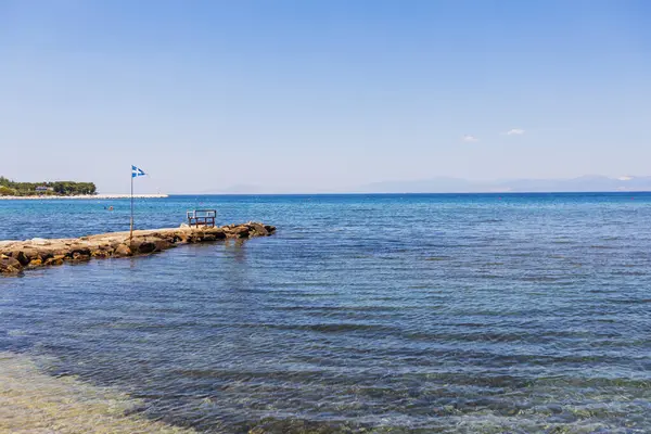 Berrak mavi suyla sakin deniz manzarası, dinlenmek için görkemli dağlara ve canlı kıyı bitkilerine karşı kurulmuş bir bank ve bayrak direğinin yer aldığı tuhaf kayalık iskele..