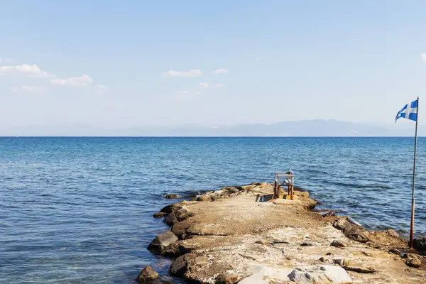Durgun denize doğru basit, kayalık iskelede dalgalanan tek bir tahta sandalye ve Yunan sahilinde gururla dalgalanan bir bayrak. Huzurlu sahne bir huzur ve yalnızlık hissi yakalıyor..