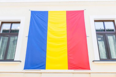 Krem rengi bir binanın dış tarafında belirgin şekilde mavi, sarı ve kırmızı dikey çizgileri bulunan üç renkli Romanya bayrağı görülüyor. Simetrik kahverengi çerçeveli pencereler arasında asılı ulusal sembol.