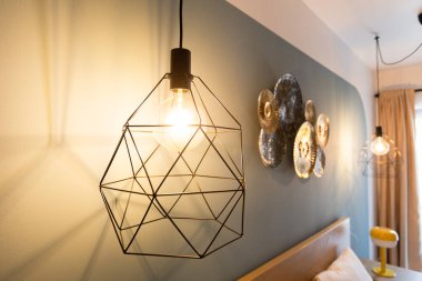 Siyah geometrik tel kafes tasarımı ve klasik Edison ampulü içeren modern endüstriyel kolye ışığı. Çağdaş aydınlatma armatürü arka planda dekoratif altın disklerle dramatik gölgeler oluşturur.