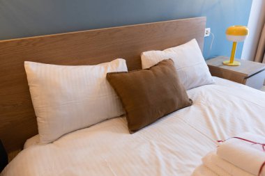 Modern otel odasının iç kısmında meşe ağacından bir yatak başlığı, gevrek beyaz yatak örtüsü ve desenli kahverengi aksanlı yastık var. Yumuşak nötr renk paleti ve ortam aydınlatmalı minimalist yatak odası tasarımı.