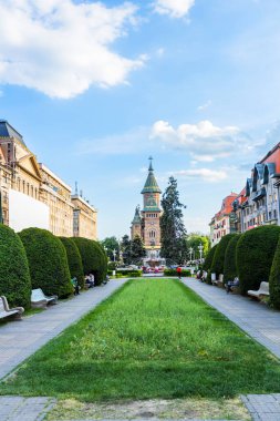 Timioara 'daki Zafer Meydanı (Piaa Victoriei) manzarası, manikürlü bahçeler, yürüyüş yolları, banklar ve mavi gökyüzünün altında kulesi olan ikonik kültürel yapı..