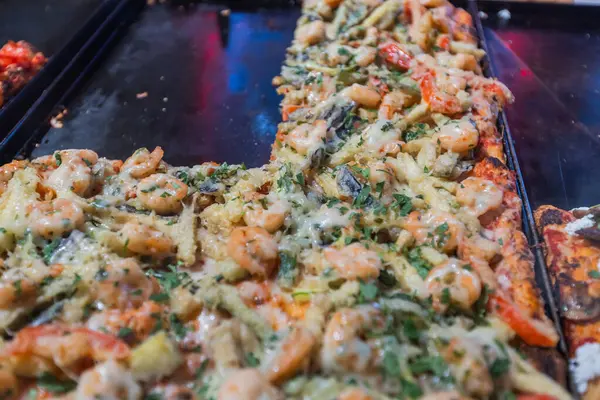 Cam tezgahın arkasındaki tepsilerde sunulan farklı malzemelerle çeşitli lezzetli pizza dilimlerinin yakın plan görüntüsü. Sunulmaya hazır ya da götürülmeye hazır cazip bir yemek seçimi vurgulanıyor.