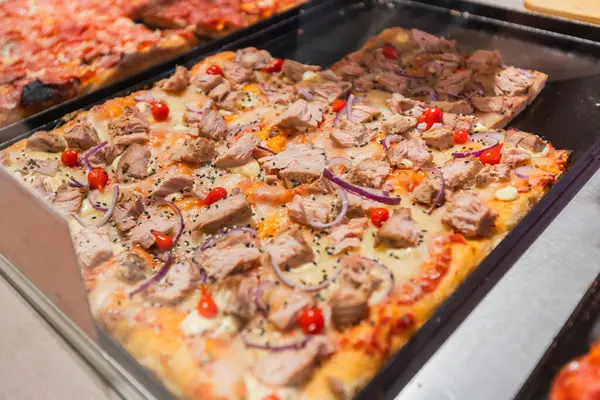 Cam tezgahın arkasındaki tepsilerde sunulan farklı malzemelerle çeşitli lezzetli pizza dilimlerinin yakın plan görüntüsü. Sunulmaya hazır ya da götürülmeye hazır cazip bir yemek seçimi vurgulanıyor.