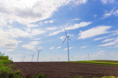 Parlak, bulutlu bir gökyüzü altında kırsal alanda temiz ve yenilenebilir enerji üreten çoklu modern rüzgar türbinlerinin manzarası..