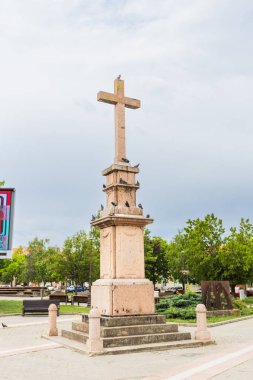 Tanınmış taş haç heykeli, Sırbistan 'ın Vrsac kentinde tüneyen güvercinlerle süslenmiş sakin bir halk parkında veya meydanda duruyor..