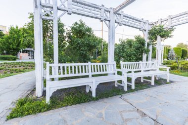 Halka açık parkta bahçe tezgahları olan beyaz ahşap Pergola. Etrafı yemyeşil ağaçlarla çevrili ve dışarıda huzurlu bir rahatlama alanı için taş yollar.