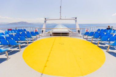 Beyaz feribot güvertesinde parlak sarı dairesel helikopter pisti. Etrafı çarpıcı okyanus ve dağ zeminiyle çevrili mavi yolcu koltuklarıyla çevrili. Mükemmel deniz taşımacılığı görüntüsü.