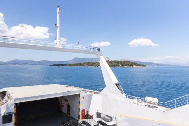 Feribot güvertesi perspektifi kristal mavi sularda beyaz gemi direği, korkuluklar ve orman adasını gösteriyor. Kabarık beyaz bulutları ve dağlık kıyı manzarası olan güneşli Akdeniz deniz manzarası.