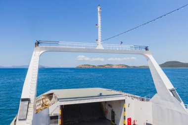 Feribotun açık yay rampasından çekilen eşsiz bir perspektif görüntüsü açık, güneşli bir gökyüzünün altındaki engin, mavi denizin uzak, tepelik adalara doğru uzandığını gösteriyor..