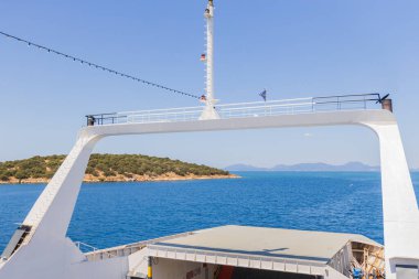 Feribotun açık yay rampasından çekilen eşsiz bir perspektif görüntüsü açık, güneşli bir gökyüzünün altındaki engin, mavi denizin uzak, tepelik adalara doğru uzandığını gösteriyor..