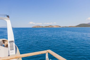 Feribotun açık yay rampasından çekilen eşsiz bir perspektif görüntüsü açık, güneşli bir gökyüzünün altındaki engin, mavi denizin uzak, tepelik adalara doğru uzandığını gösteriyor..