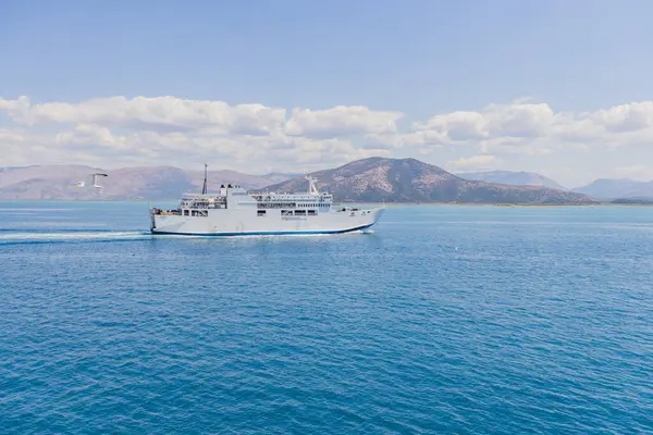 Beyaz yolcu feribotu kristal berrak turkuaz turkuaz Akdeniz sularından geçiyor dramatik Yunan adaları ve dağ manzarası parlak mavi gökyüzünün altında görülebiliyor..