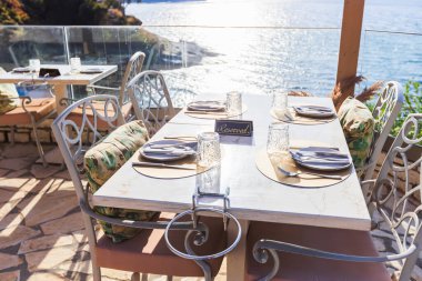 Güneşli bir terasta panoramik deniz manzaralı manzaralı açık hava yemek masası. Örülmüş sandalyeler ve minimalist sofra takımı rahat ama rafine bir atmosfer yaratıyor, tatil ve konukseverlik için ideal..