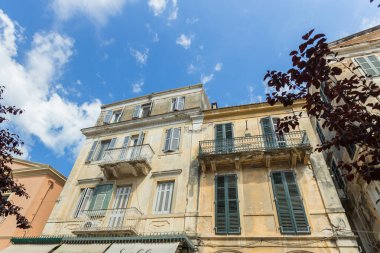 Corfu Town 'daki klasik bir Yunan binasının şaşaalı manzarası, süslü balkonların ve karmaşık mimari detayların yer aldığı. Yapı güneş ışığıyla yıkanmış, açık mavi gökyüzüne karşı kurulmuş, Akdeniz cazibesinin özünü yakalıyor..