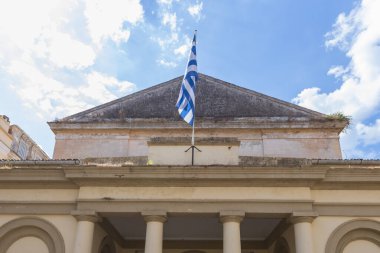 Yunanistan 'ın Korfu kentindeki tarihi Eski Belediye Binası' nın düşük açılı manzarası, klasik sütunlar ve üçgen bir pedikürle, Yunan bayrağı binanın tepesinde gururla dalgalanırken.