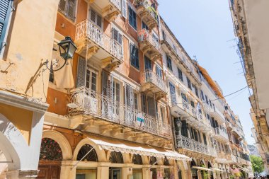 Korfu Eski Kasabası 'nda güneş gören geniş bir cadde, birden fazla katlı geleneksel Venedik tarzı binalar, dekore edilmiş demir balkonlar, Avrupa sokak manzarası.