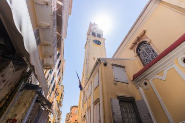 Aziz Spyridon Kilisesi 'nin ikonik çan kulesinin düşük açılı görüntüsü, belirgin kırmızı kubbesi ve mavi saat yüzüyle, güneşli bir günde Corfu Eski Kasabası' ndaki dar, geleneksel Venedik tarzı binaların üzerinde yükseliyor. Yunanistan.