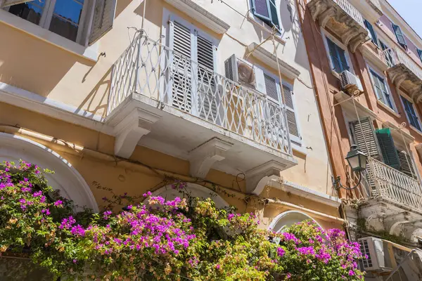 Korfu 'daki geleneksel bir binanın detaylı bir görüntüsü, beyaz dövülmüş demir bir balkona odaklanmış ve zemin kattaki kemerlerin üzerinde çağlayan pembe begonviller..