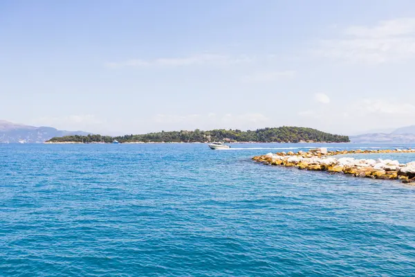 Turkuaz denizi, kayalık rıhtımı ve yemyeşil bir adaya karşı sürat yapan bir motorlu tekne ile güneşli mavi gökyüzünün altında.