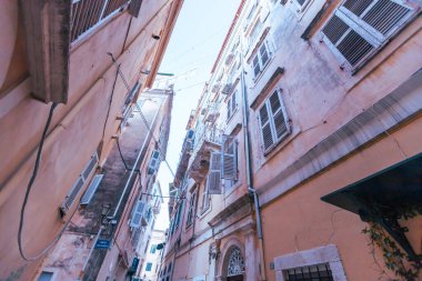 Dar kentsel alanı ve gökyüzünü aydınlatan, iki eski pembe-bej bina arasındaki dar bir ara sokağa bakan dramatik bir alçak açılı çekim. Corfy, Yunanistan.