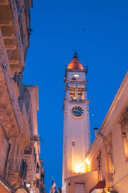 Görkemli St. Spyridon çan kulesi aydınlanmış saat yüzüyle Corfu 'nun eski kasabasındaki büyülü mavi saatte geleneksel Yunan mimarisinin üzerinde yükseliyor..