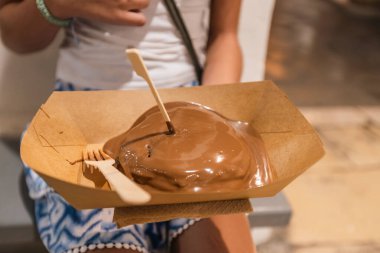 Yakından çekilmiş bir fotoğraf, elinde karton tepsiyle yumuşak, erimiş çikolataya batırılmış ve küçük bir ahşap çatalla servis edilen bir yığın krep..