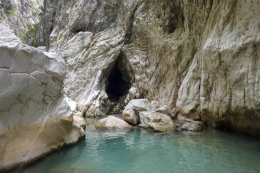 Duvarında delik olan küçük bir mağara. Su berrak ve mavi. Suda bir kaya var.