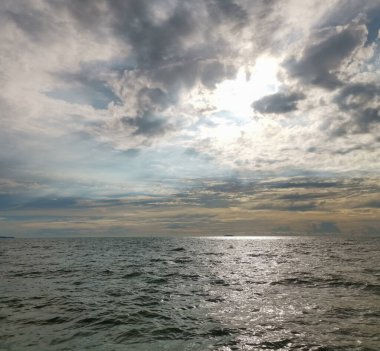 Gökyüzü bulutlu ve güneş bulutların arasında parlıyor. Okyanus sakin ve su güneşi yansıtıyor.