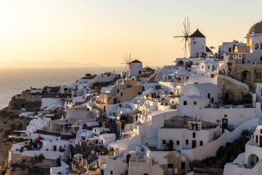 Oia, Santorini, Yunanistan - 28 Haziran 2021: Oia 'da, Santorini Adası' nda günbatımının ılık ışınlarında beyazlatılmış evler ve yel değirmenleri. Yunanistan