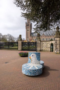 The Hague, Netherlands - April 17, 2023: A bench with a white dove holding an olive branch a symbol of peace in front Peace Palace in The Hague, which houses the International Court of Justice clipart
