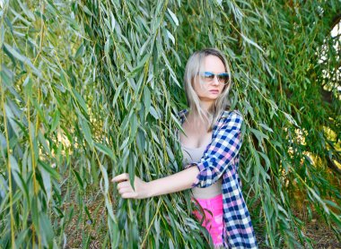 Güneş gözlüklü güzel bir kadın yazın söğüt ağacının kalın, yeşil dallarına yaslanır..