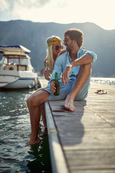 大人の白人カップルはドックに座って笑顔で笑ってビールを飲み海辺で楽しい時間を過ごしていました — ストック写真