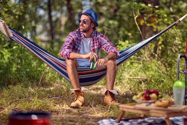 Młody Przystojny Hipsterski Mężczyzna Siedzący Hamaku Trzymający Piwo Piknik Lesie — Zdjęcie stockowe