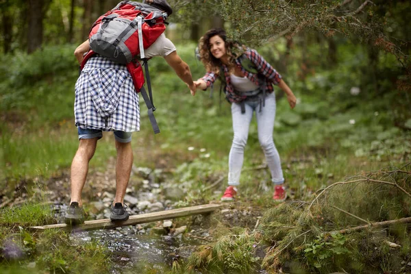 ボーイフレンドは 彼らが自然の中で美しい一日にハイキング中に小川を渡るために彼のガールフレンドを助けています ハイキング アクティビティ — ストック写真