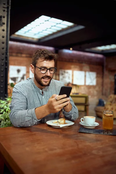 戴眼镜的男人坐在咖啡店里 在手机上打滚 喝咖啡休息 生活方式 麻风病概念 — 图库照片