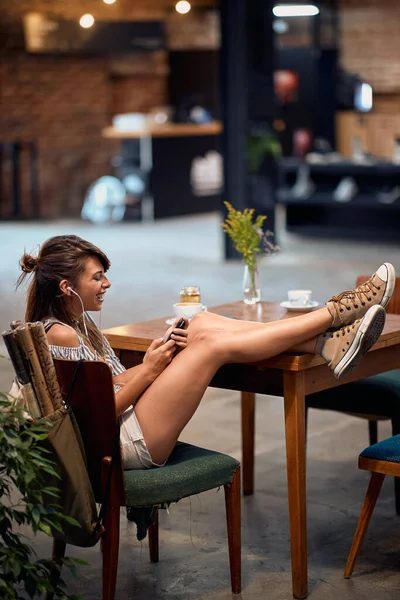 イヤホン付きの美しい若いヒップスターの女の子は 単独で足をカフェで喜びを感じてテーブルに座っています ライフスタイル 学生のコンセプト — ストック写真