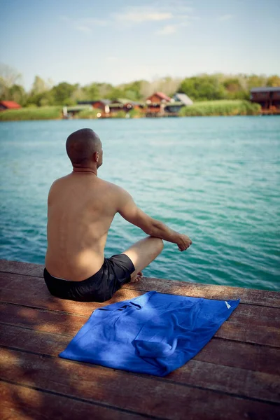 Man doing morning meditation by water. Young man sitting on wooden jetty by sea doing spiritual practice. Relaxation, discipline, lifestyle concept.