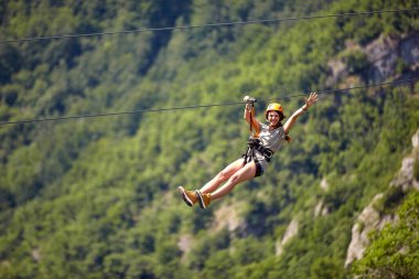 Ormanda halatla kayarken günlük giysiler giyen neşeli, genç bir bayan turist. Bulanık arka plana karşı halatla kayma gezisi seçici odak. Tatil, macera, ekstrem spor konsepti.