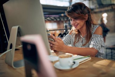 Modern yaratıcı bir ofiste akıllı telefon kullanarak masaüstü bilgisayarının önünde çalışan güzel bir genç kadın. İş, çalışan, öğrenci konsepti.