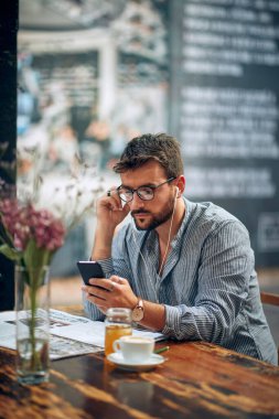 Gözlüklü yakışıklı genç adam kulaklıklı akıllı telefon kullanıyor, tek başına kahve molasının tadını çıkarıyor. Modern kafe içi.