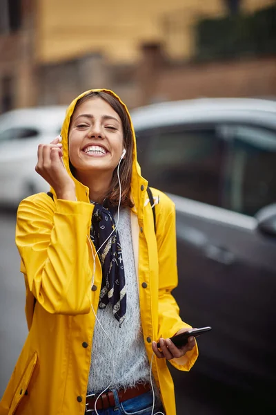 一个穿着黄色雨衣的年轻女孩一边欣赏着音乐 一边在雨中平静地穿过城市 一边摆姿势拍照 — 图库照片