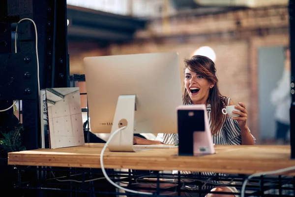 Радостная Молодая Женщина Смотрит Экран Держа Кофе Сидя Офисе Чувствуя — стоковое фото