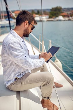 Genç bir adam sıcak güneşin altında, rahat bir atmosferde yatta oturuyor ve tablet kullanıyor. Yaz, deniz, tatil