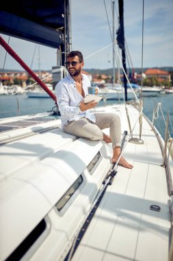 Tabletli genç bir adam yatta oturuyor ve deniz kenarında güneşli bir günde rıhtımın güzel manzarasının ve kahvenin tadını çıkarıyor. Yaz, deniz, tatil
