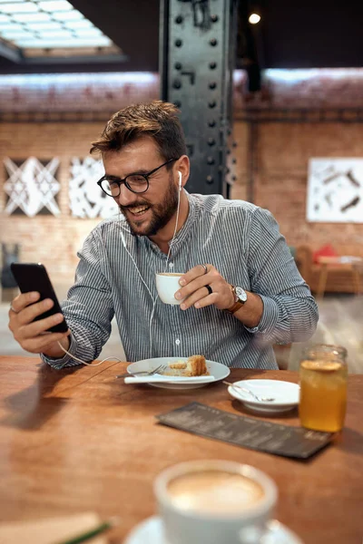 餐厅里一个英俊快乐的男人看着电话 拿着并享受着一杯咖啡 在电话里打滚 — 图库照片