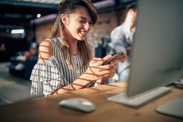 スマートフォンを使用してデスクトップコンピュータの前にテーブルに座っている美しい若い女性は 喜びを感じている ビジネス オフィスの仕事の概念 — ストック写真